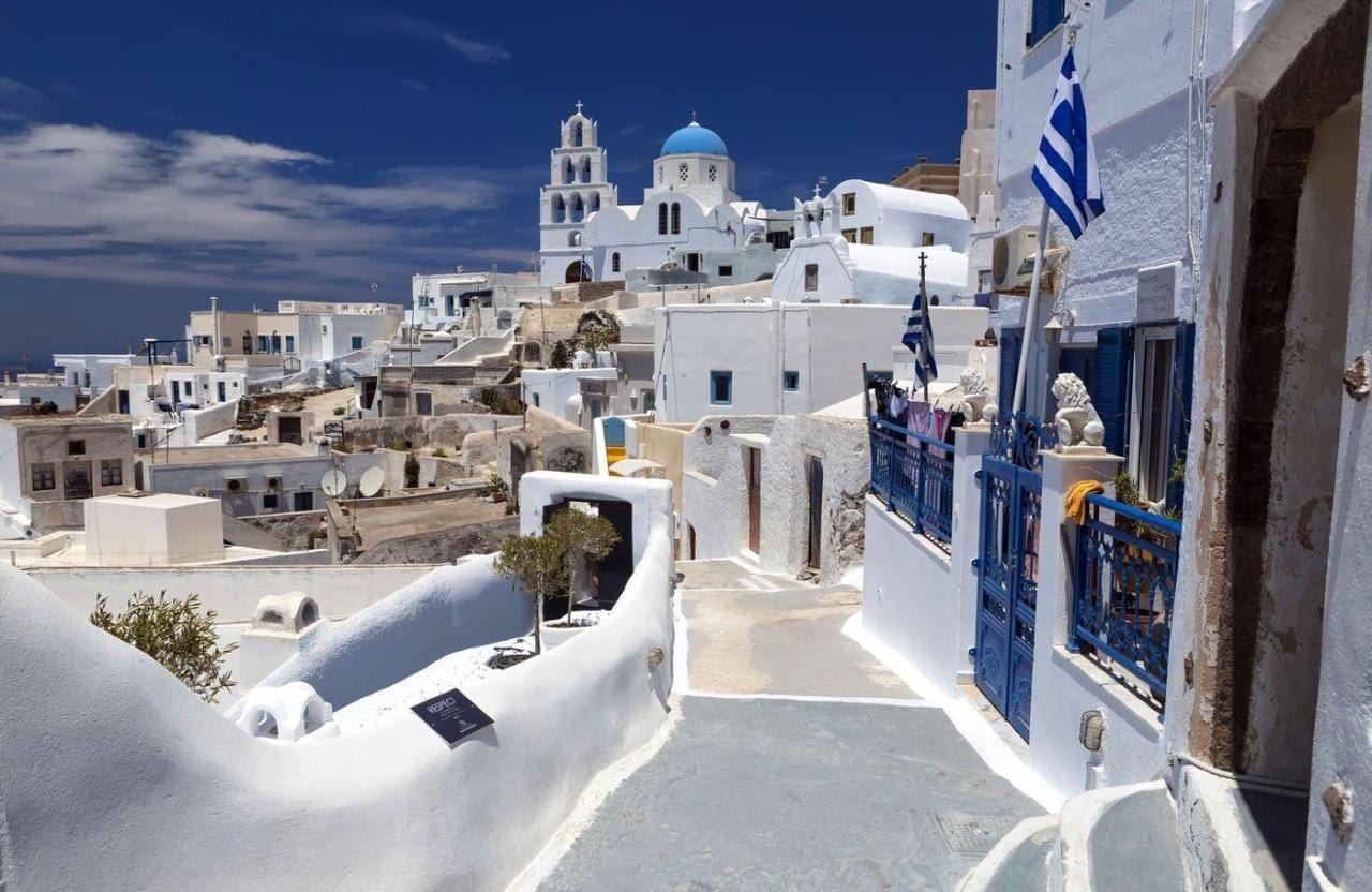 A blue-domed church and Greek flag above a whitewashed lane in Pyrgos